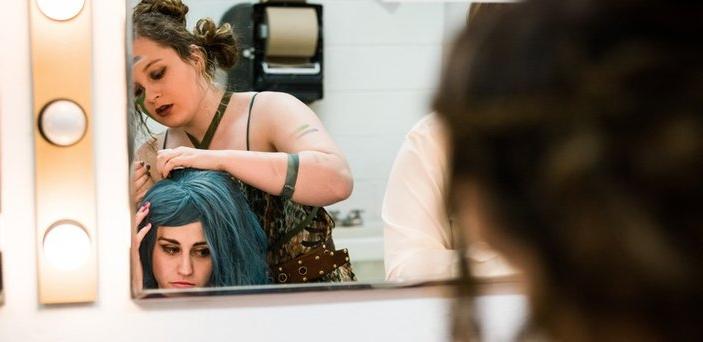 One actress helping another actress with her wig in a mirror backstage